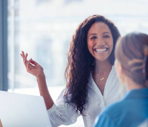 Business colleagues having a conversation. They are both young business people casually dressed in a modern office. Could be an interview or consultant working with a client. She is listening and smiling. One person has her back to us. Mixed ethnic group. One is African American and the other is Caucasian